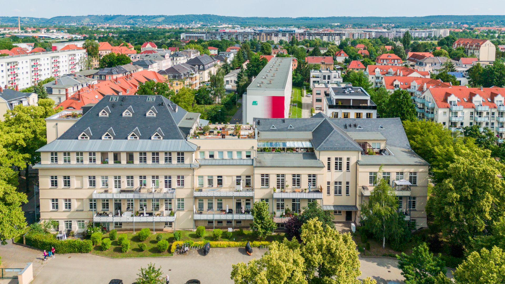Großes helles Wohngebäude mit mehreren Etagen, Bäumen und Garten im Vordergrund.