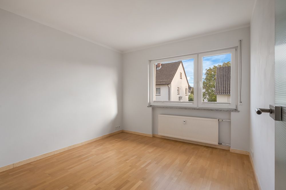 Helles, leeres Zimmer mit Parkettboden, großem Fenster und weißer Wand.