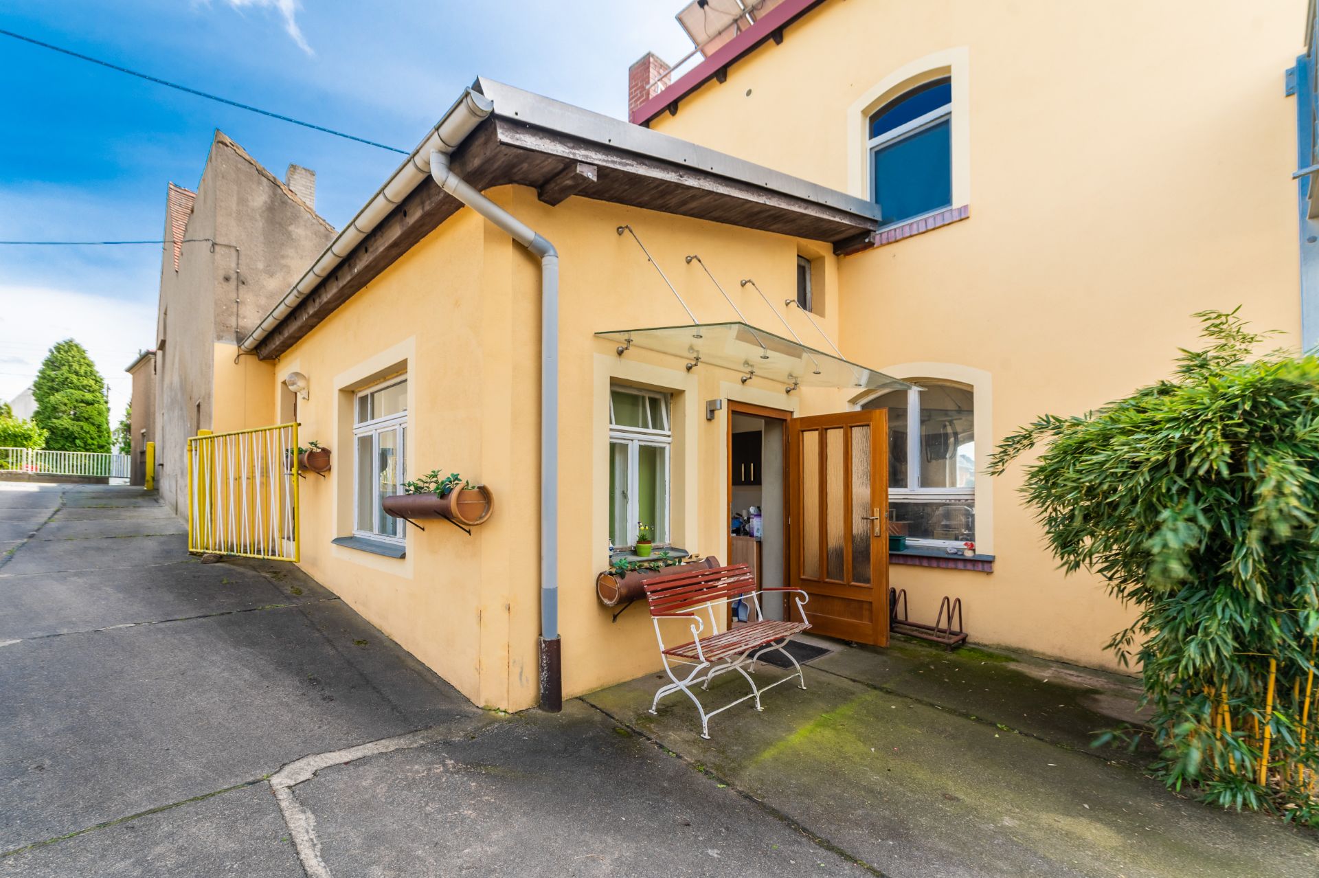 Gelbes Wohnhaus mit überdachtem Eingang, Bank und Pflanzkübeln an den Fenstern.