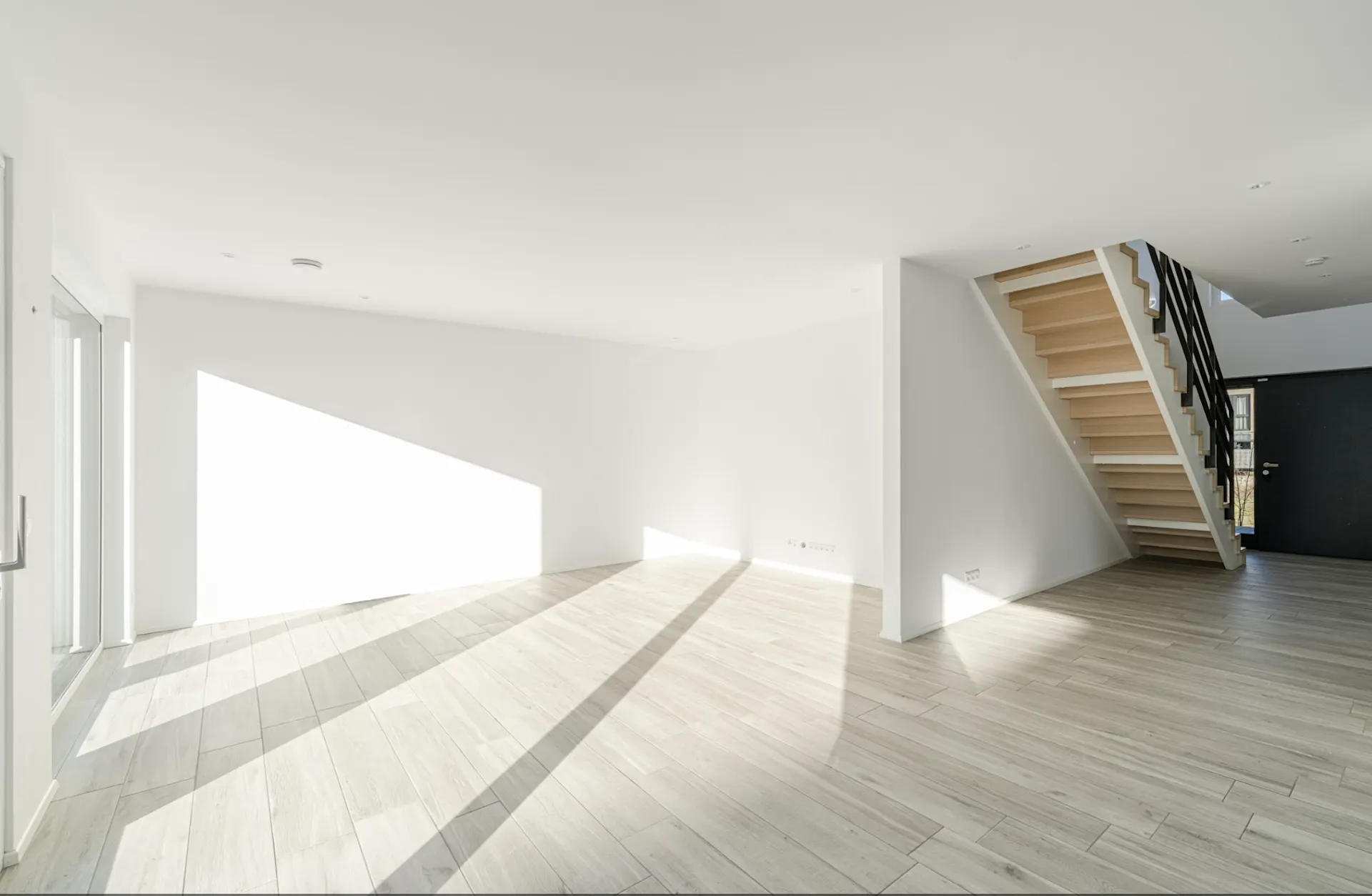 Heller, leerer Raum mit Holztreppe und großen Fenstern, Licht fällt auf den Boden.