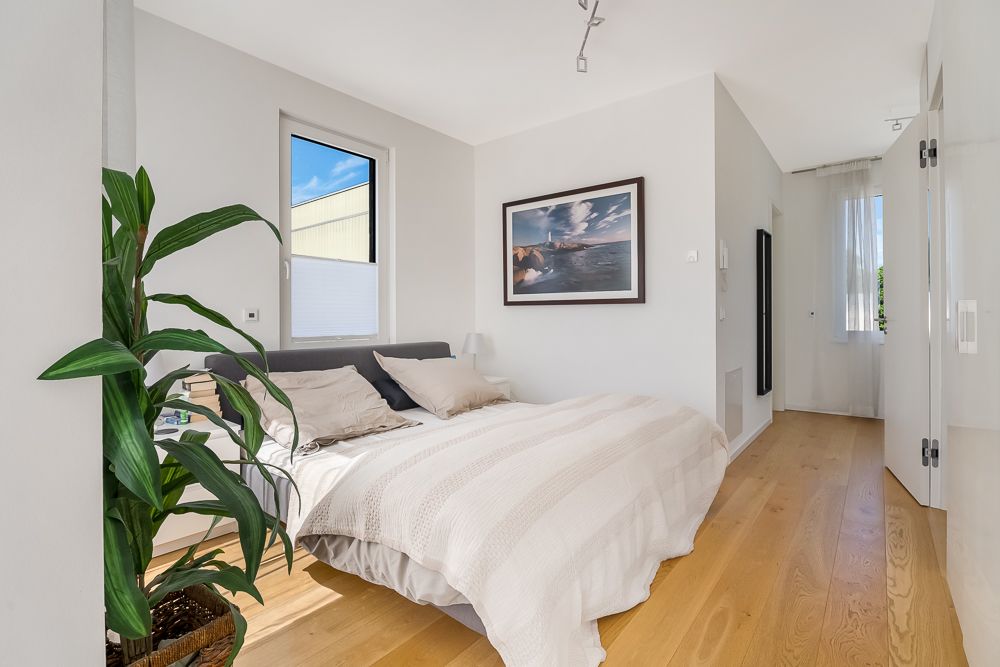 Helles Schlafzimmer mit Doppelbett, Holzboden, Pflanze und großem Fenster.