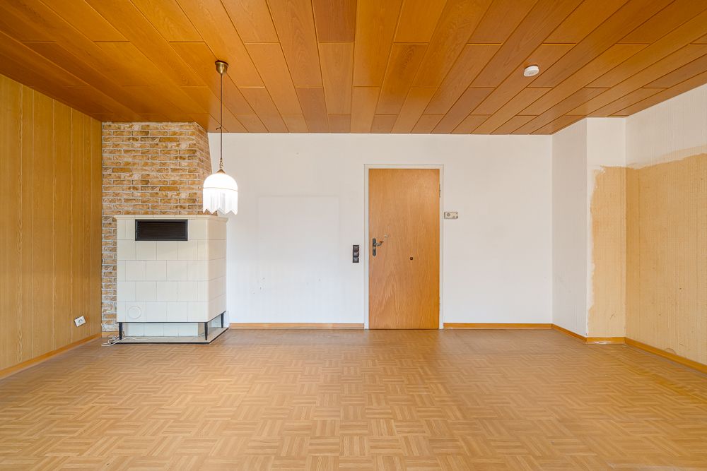 Leeres Zimmer mit Holzboden, Holzdecke, Tür, Ofen und Hängelampe an der Decke.