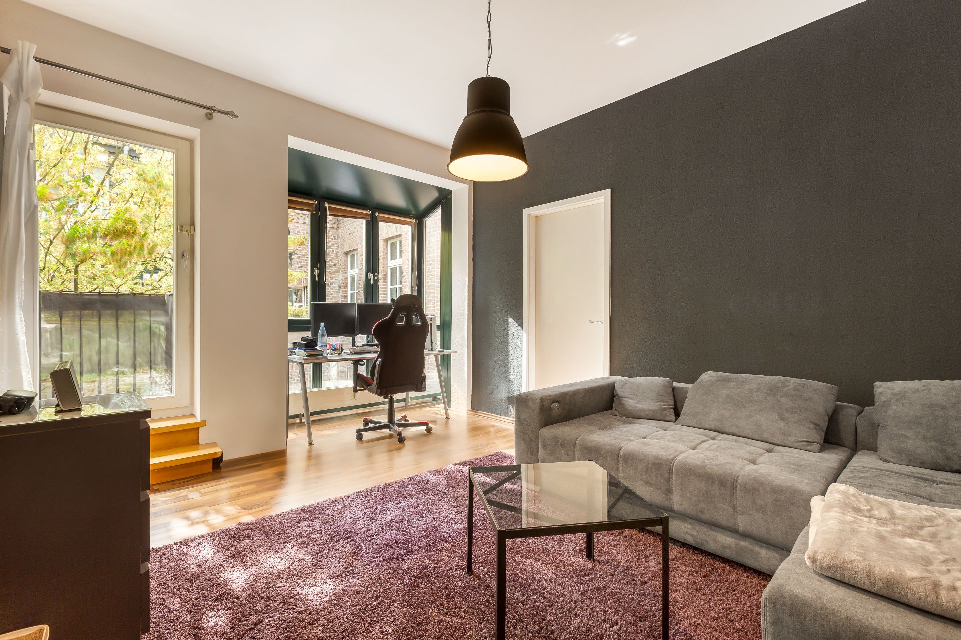 Modernes Wohnzimmer mit grauem Sofa, Glastisch, Teppich und Arbeitsplatz am Fenster.