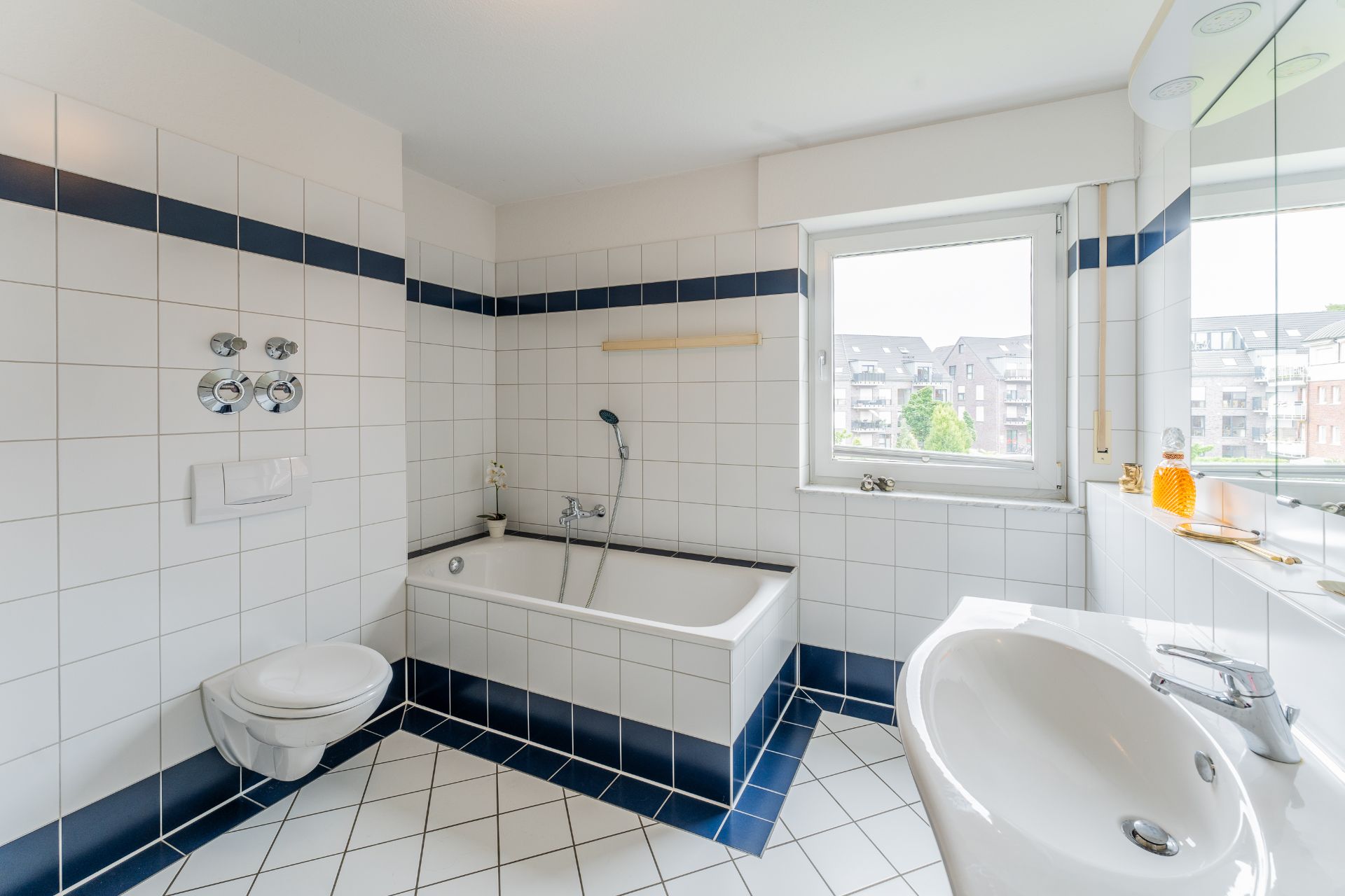 Helles Badezimmer mit Badewanne, Wandtoilette, Waschbecken und Fenster, blau-weiße Fliesen.