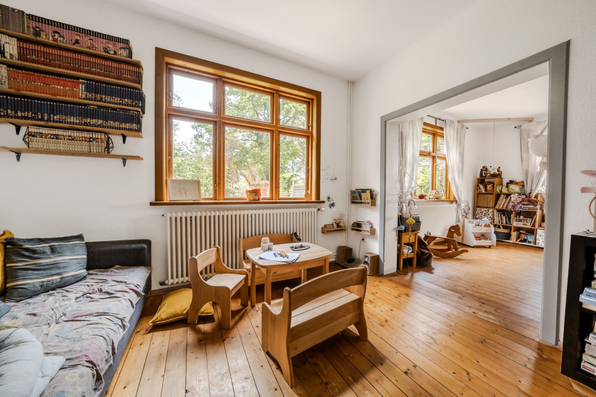 Helles Wohnzimmer mit Holzboden, Sofa, Kinderstuhlset und Bücherregalen am Fenster.