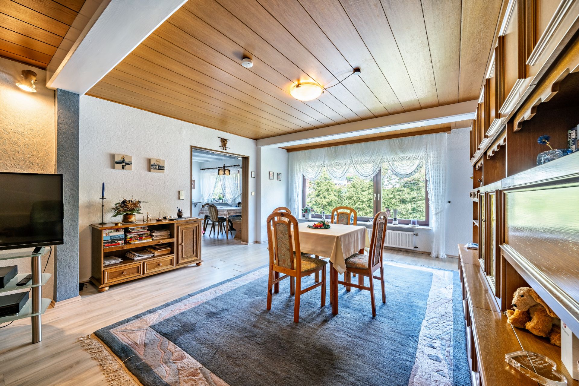 Helles Wohnzimmer mit Esstisch, vier Stühlen, großem Fenster und Holzdecke.