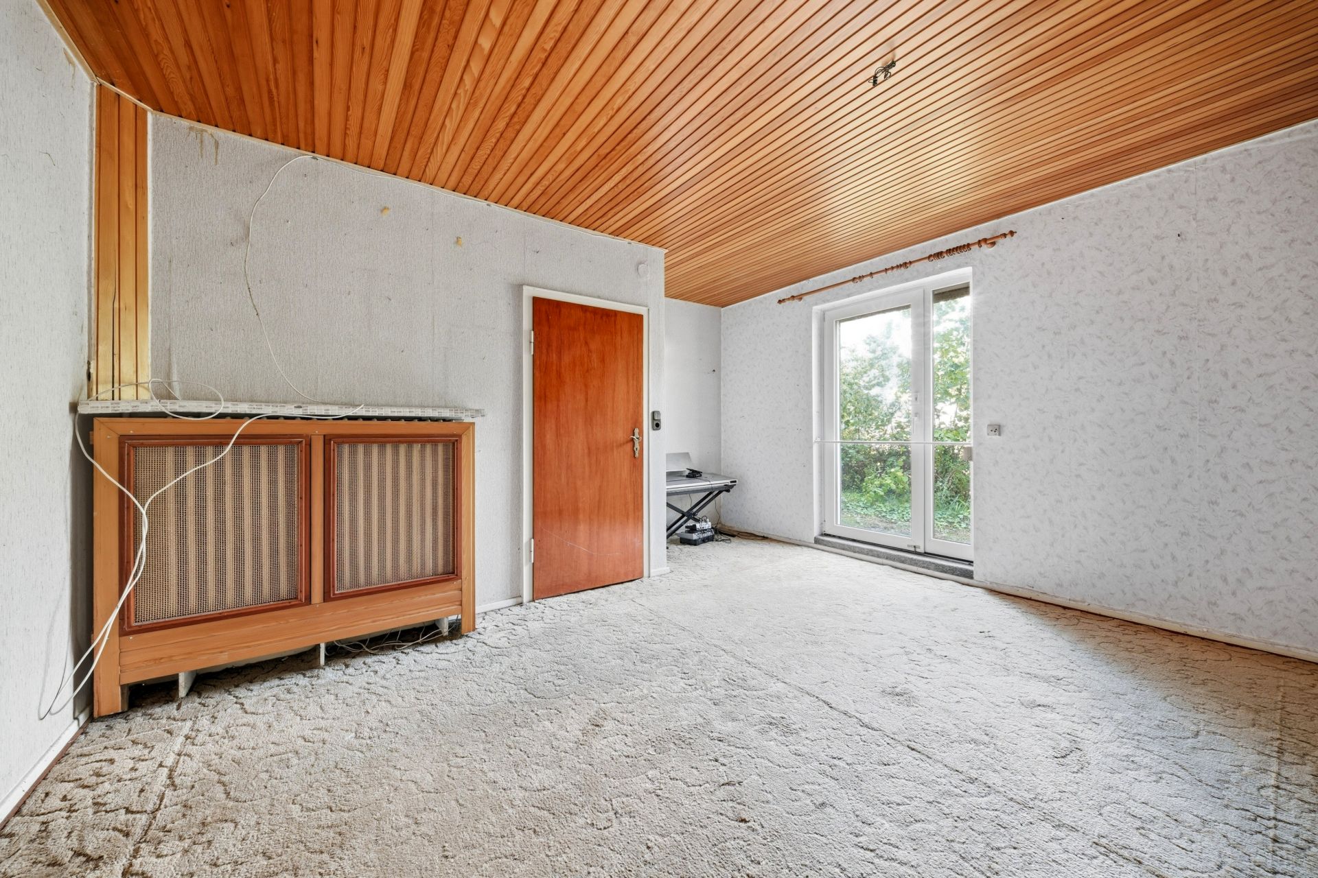Leeres Zimmer mit Holzdecke, Teppichboden, Holztür und großen Fenstertüren zum Garten.