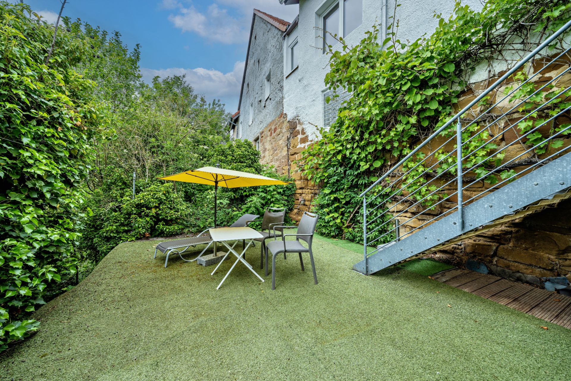 Gartenbereich mit Tisch, Stühlen, Liege und gelbem Sonnenschirm neben einer Treppe.