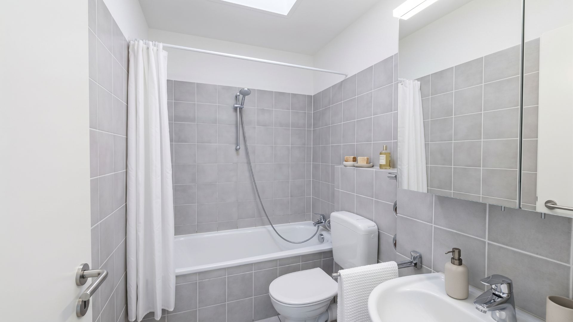 Helles Badezimmer mit Badewanne, Dusche, WC und Waschbecken, graue Fliesen und Spiegel.