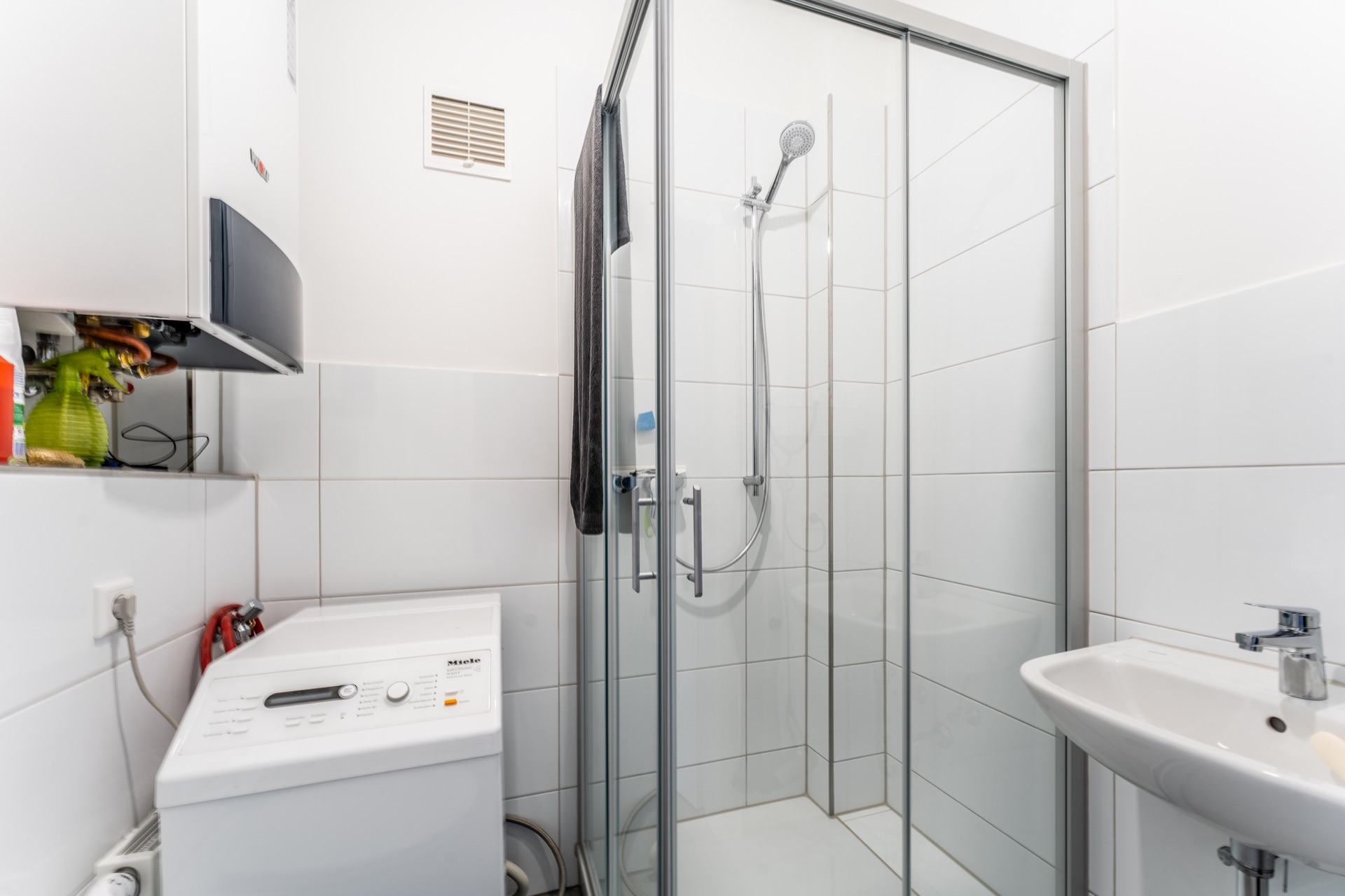 Kleines Bad mit Dusche, Waschmaschine, Waschbecken und Heizgerät an der Wand.