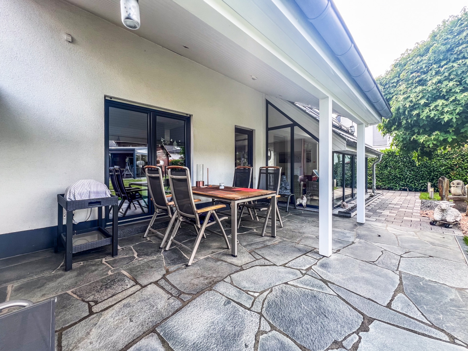 Überdachte Terrasse mit Esstisch und Stühlen auf Steinboden neben Haus.