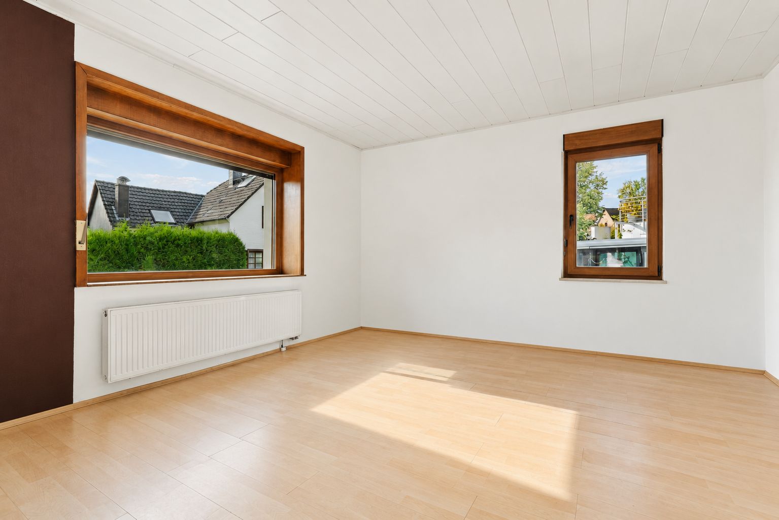 Helles, leeres Zimmer mit Holzfußboden und zwei Fenstern, weiße Wände und Decke.