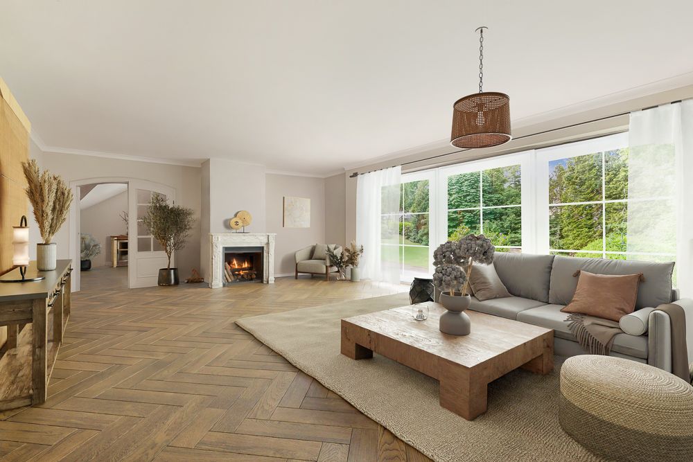Helles Wohnzimmer mit Kamin, Sofa, Couchtisch und großen Fenstern mit Blick ins Grüne.
