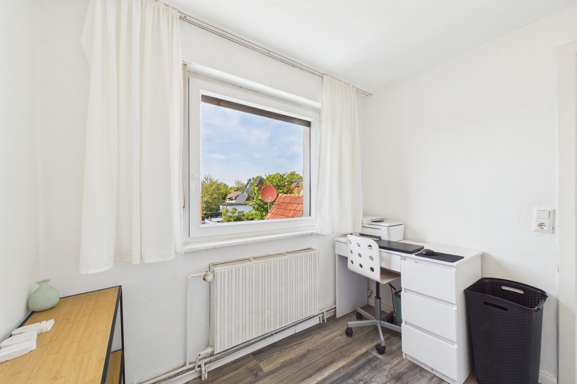 Kleines Arbeitszimmer mit Schreibtisch, Stuhl, Fenster und weißem Vorhang.