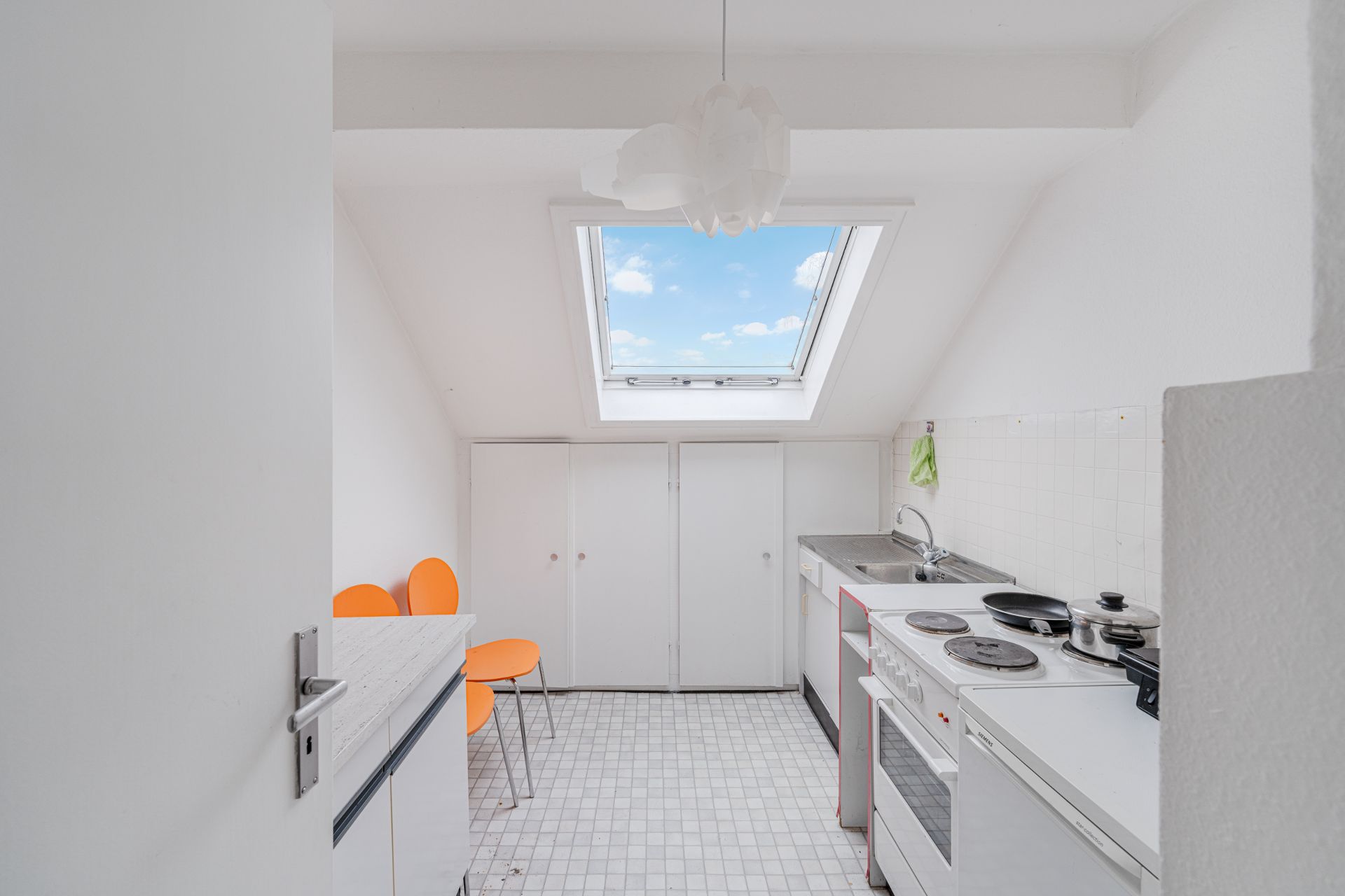 Kleine helle Küche mit Herd, Spüle, weißen Schränken, zwei orange Stühlen und Dachfenster.