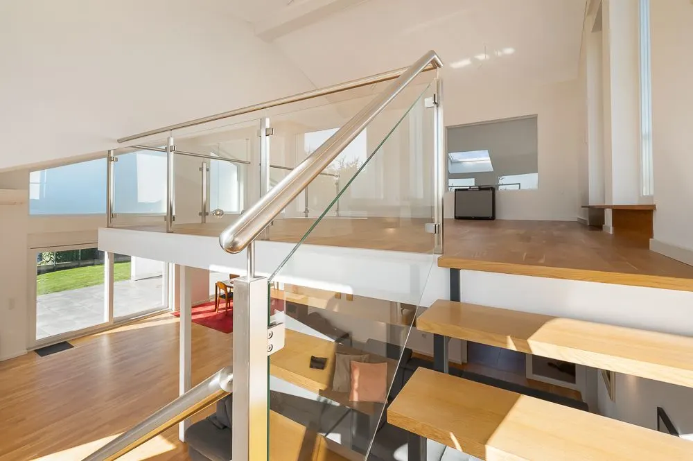 Helle, moderne Holztreppe mit Glasgeländer in einem Wohnraum mit großen Fenstern.