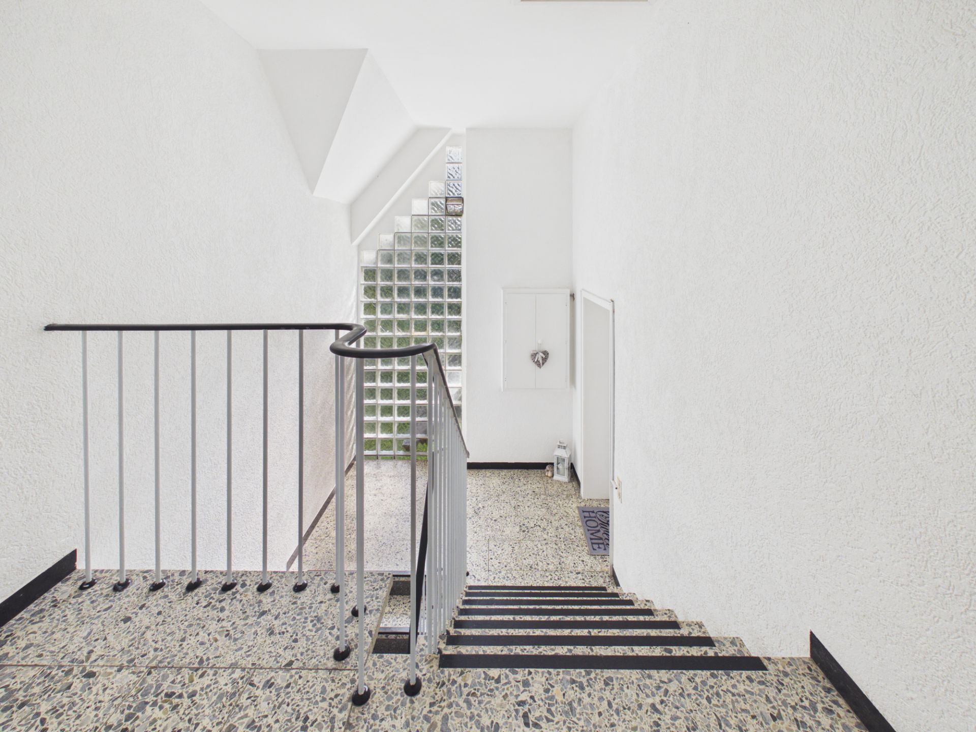 Helles Treppenhaus mit Terrazzo-Boden, Metallgeländer und Glasbausteinfenster.