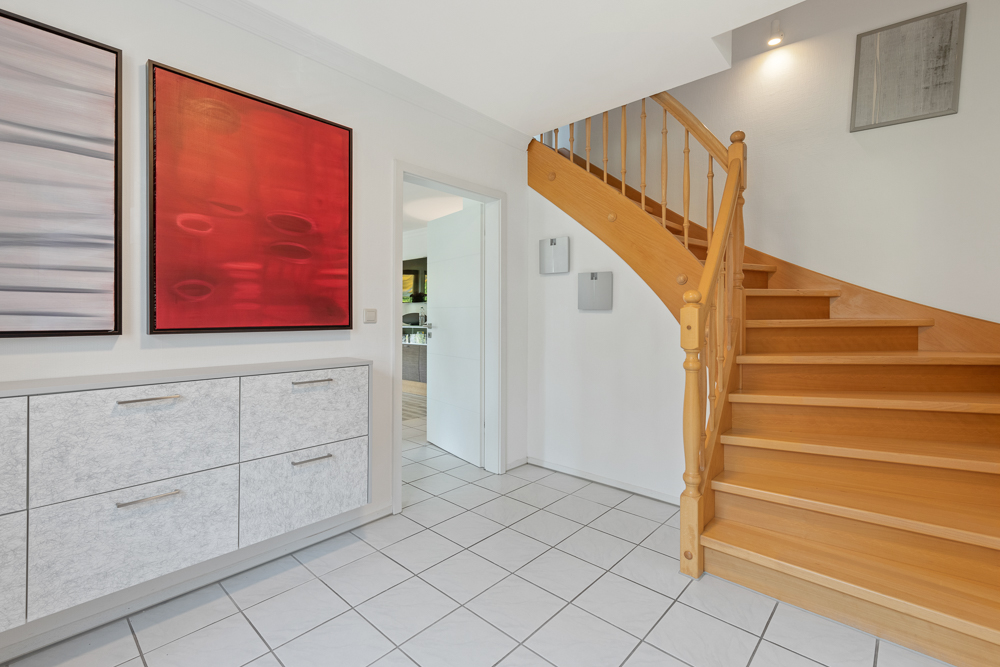 Heller Flur mit Holztreppe, weißen Fliesen, Kommode und zwei bunten Bildern an der Wand.