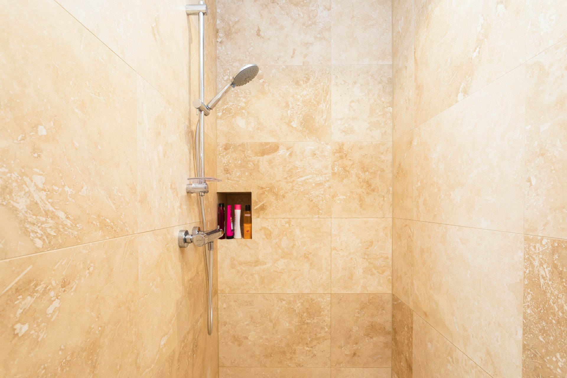 Dusche mit beigen Fliesen, Duschkopf und eingelassener Nische mit Pflegeprodukten.