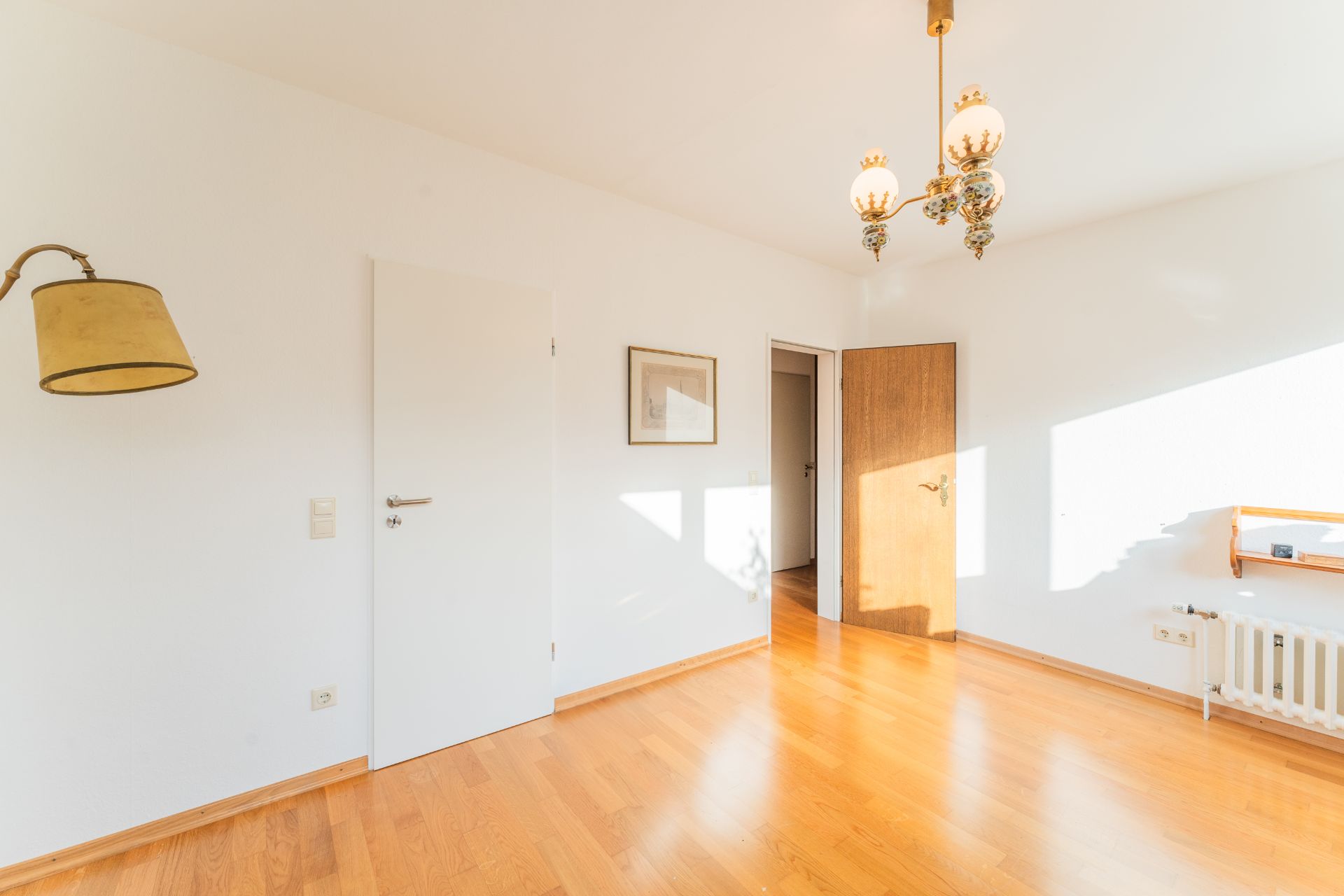 Helles, leeres Zimmer mit Holzboden, weißen Wänden, Wand- und Deckenlampen sowie zwei Türen.