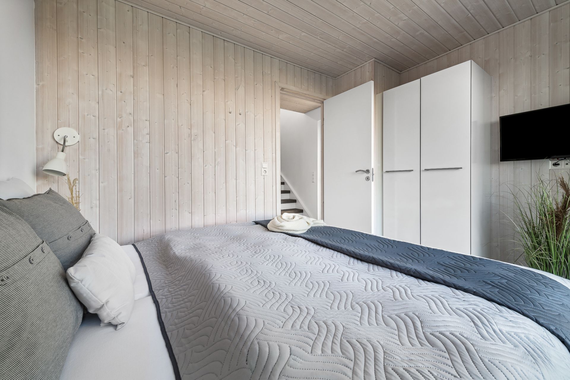 Schlafzimmer mit Doppelbett, weißem Kleiderschrank und Wandfernseher, Holzwände und Zimmerpflanze.