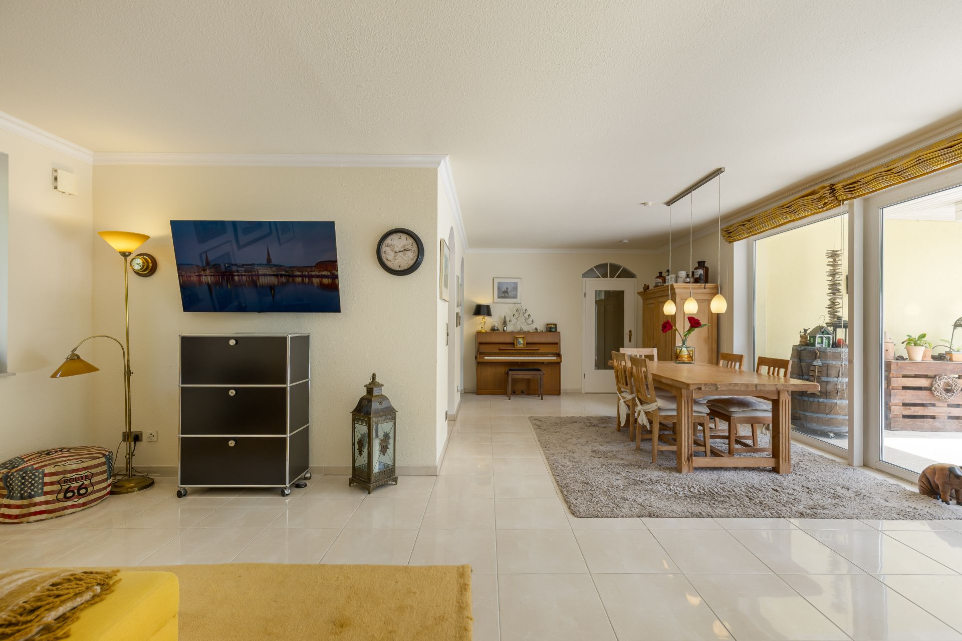 Wohnzimmer mit Esstisch, Stühlen, Fernseher an der Wand und großen Fenstern zur Terrasse.