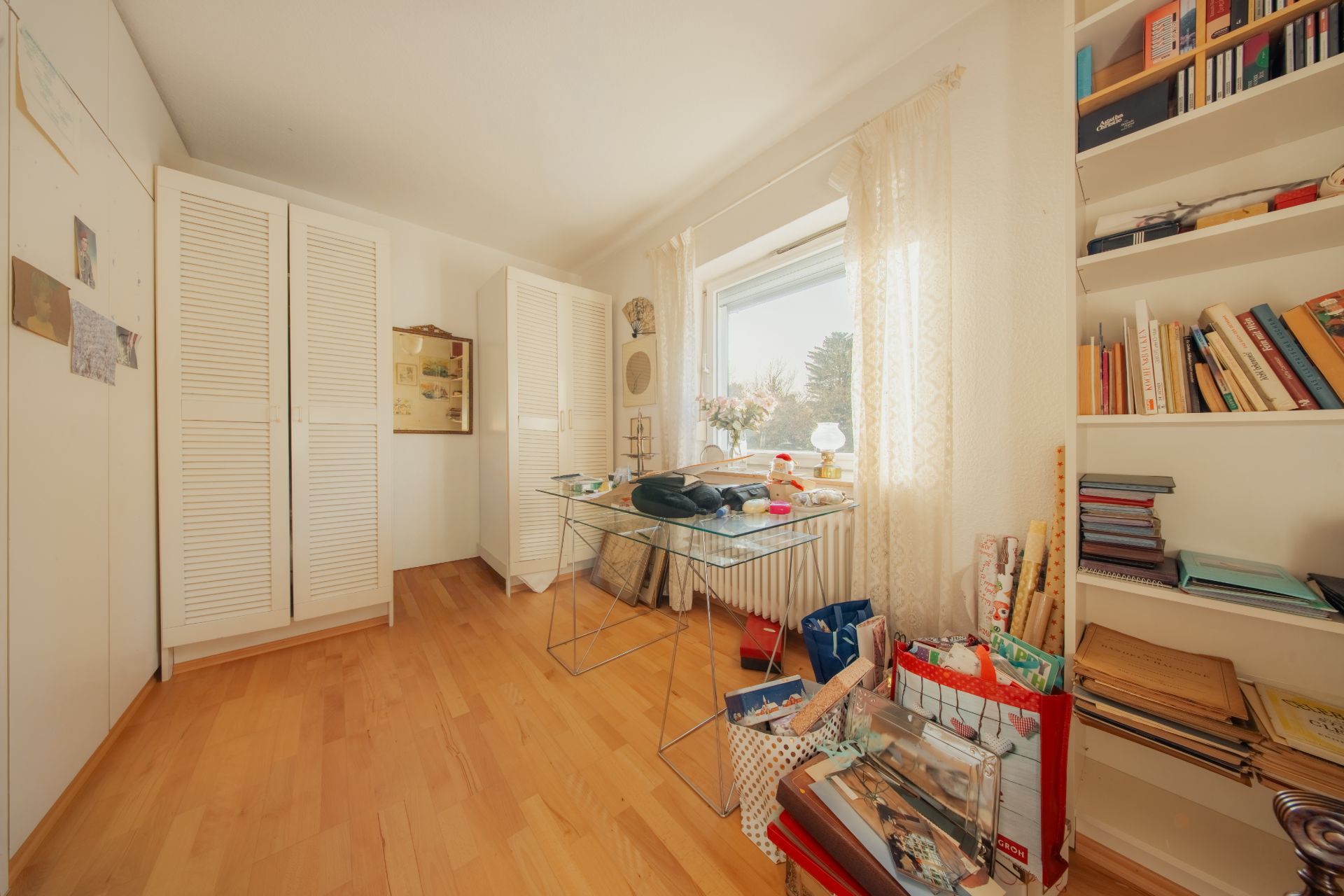 Arbeitszimmer mit Schreibtisch, Regalen, Kleiderschränken und großem Fenster mit Tageslicht.