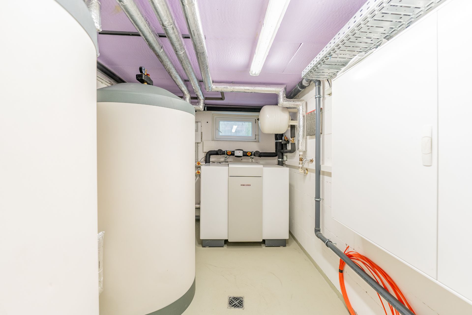 Helles Technikraum mit Heizungsanlage, Speicherbehältern und sichtbaren Rohren an der Decke.