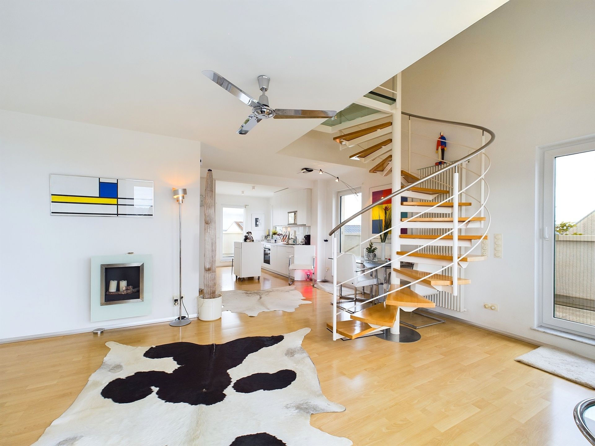 Modernes Wohnzimmer mit Holzspiraltreppe, großem Fenster, Deckenventilator und Kuhfell-Teppich.
