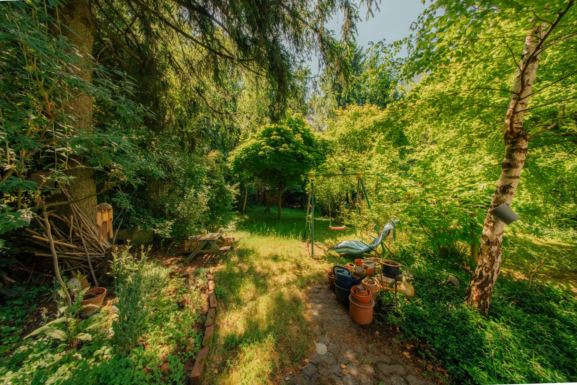 Grüner Garten mit Bäumen, Rasenfläche, Schaukel und Blumentöpfen unter hellem Sonnenlicht.