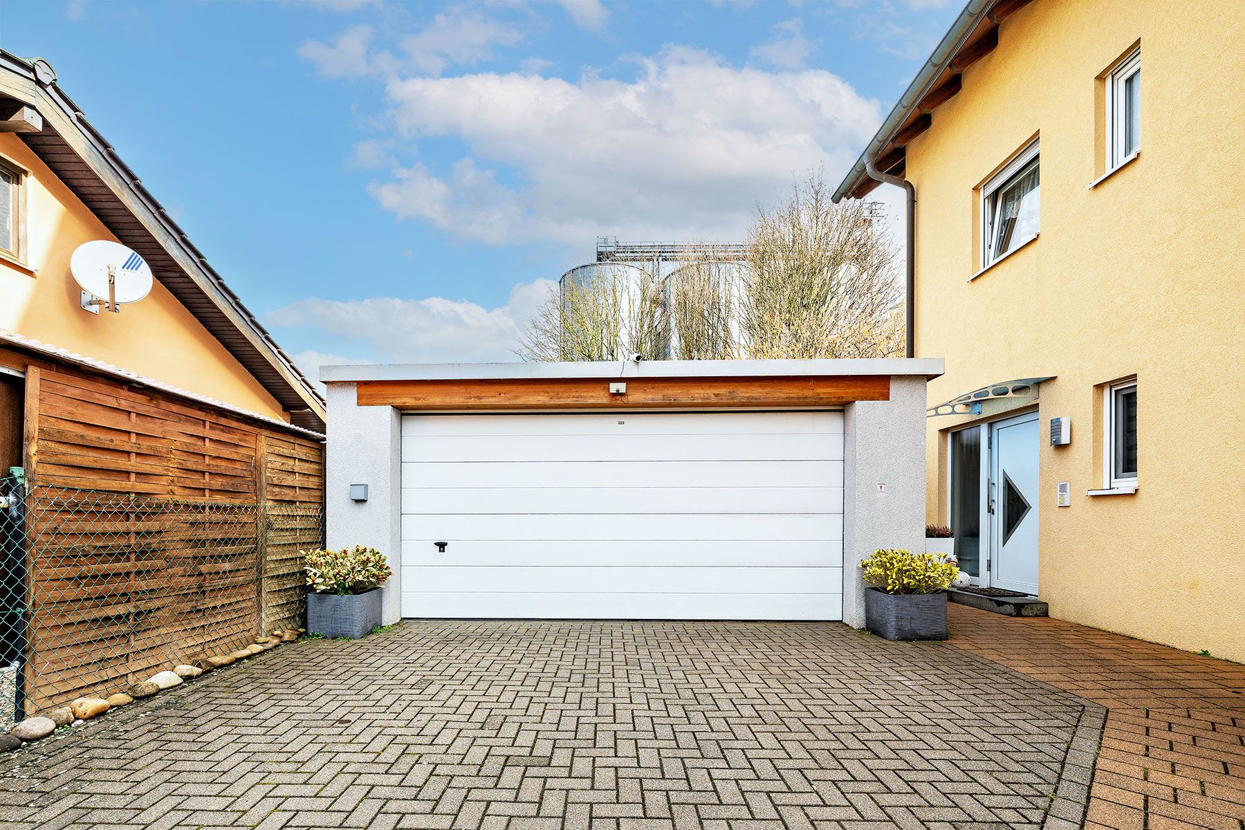 Weiße Garage mit gepflasterter Einfahrt zwischen zwei Wohnhäusern, Pflanzenkübel links und rechts.