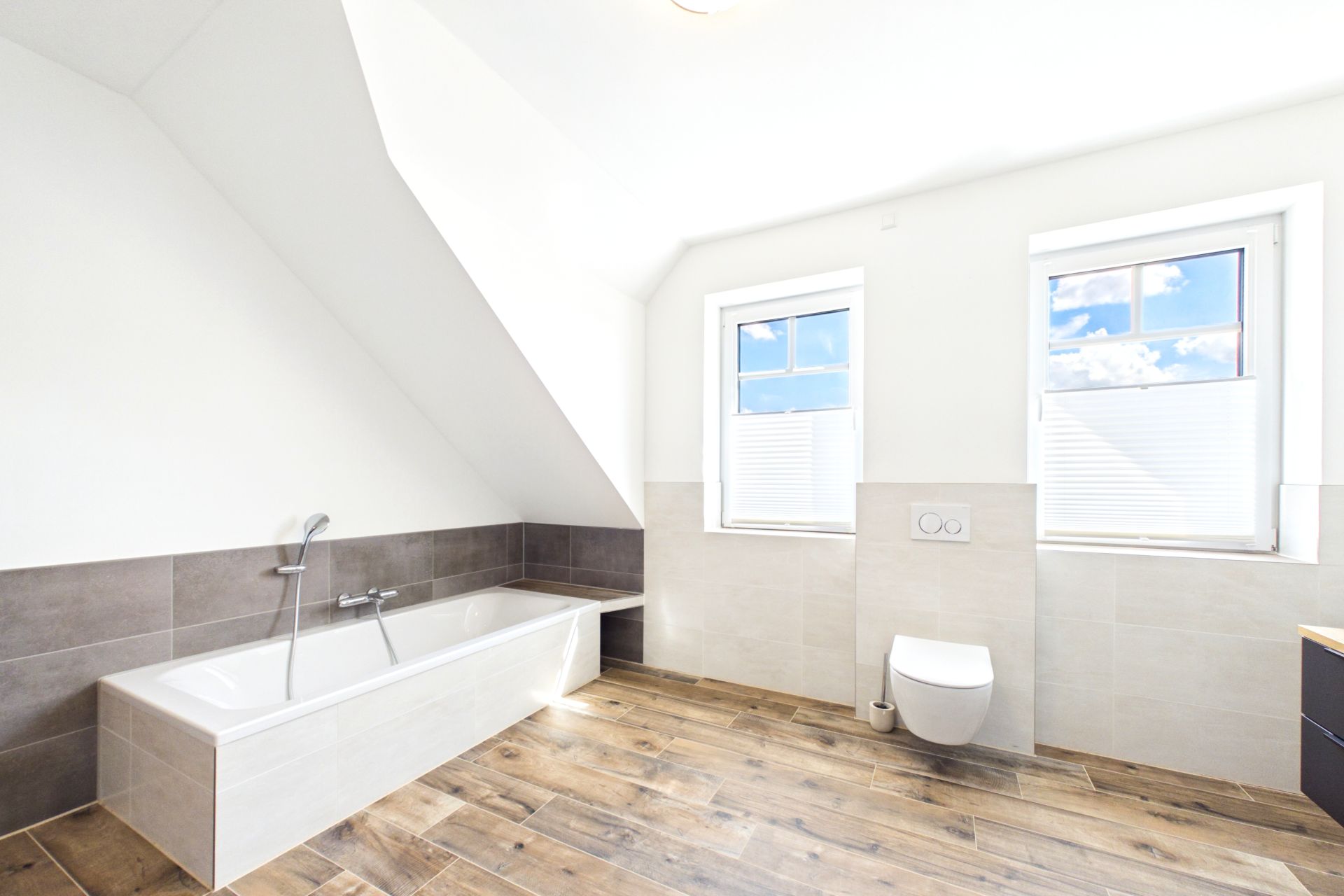 Helles Badezimmer mit Badewanne, Toilette, zwei Fenstern und Holzoptik-Fliesenboden.