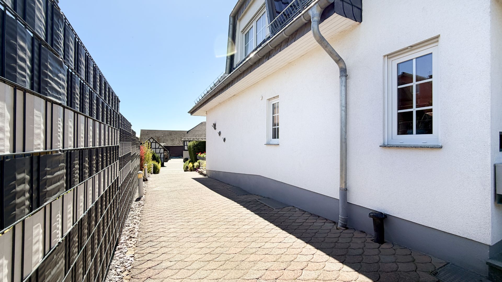 Gepflasterter Weg entlang eines weißen Wohnhauses mit Zaun und Fenstern.