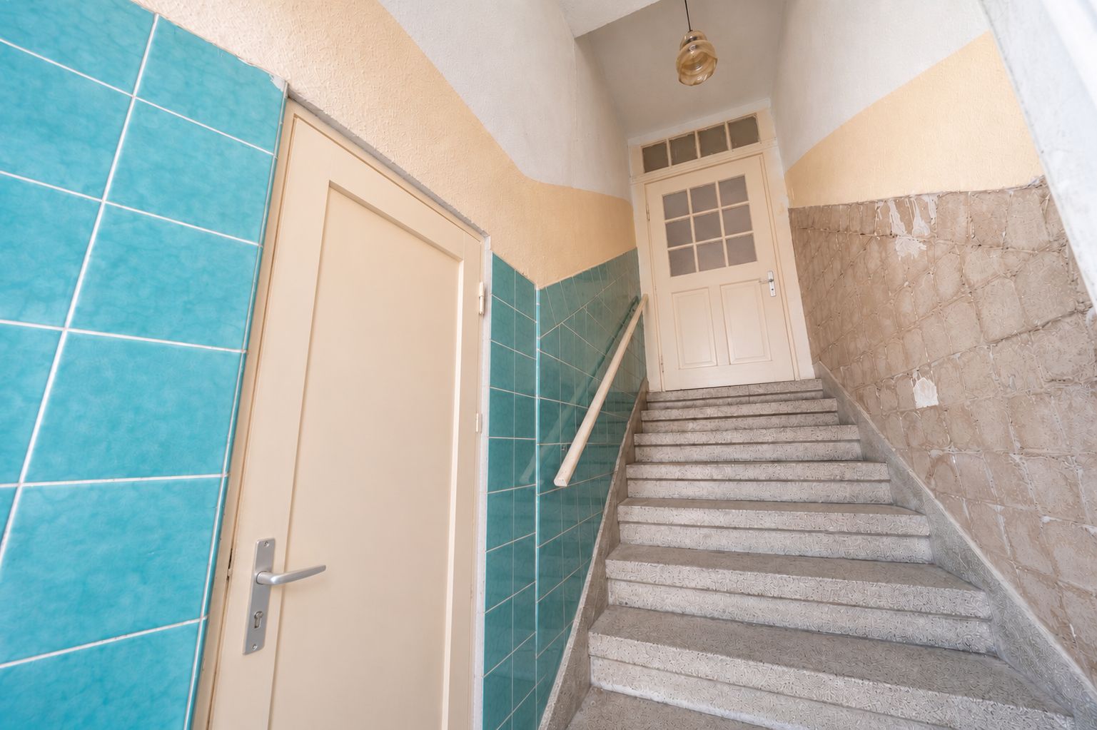 Treppenhaus mit türkisfarbenen Fliesen, einer Tür links und einer Tür oben an der Treppe.