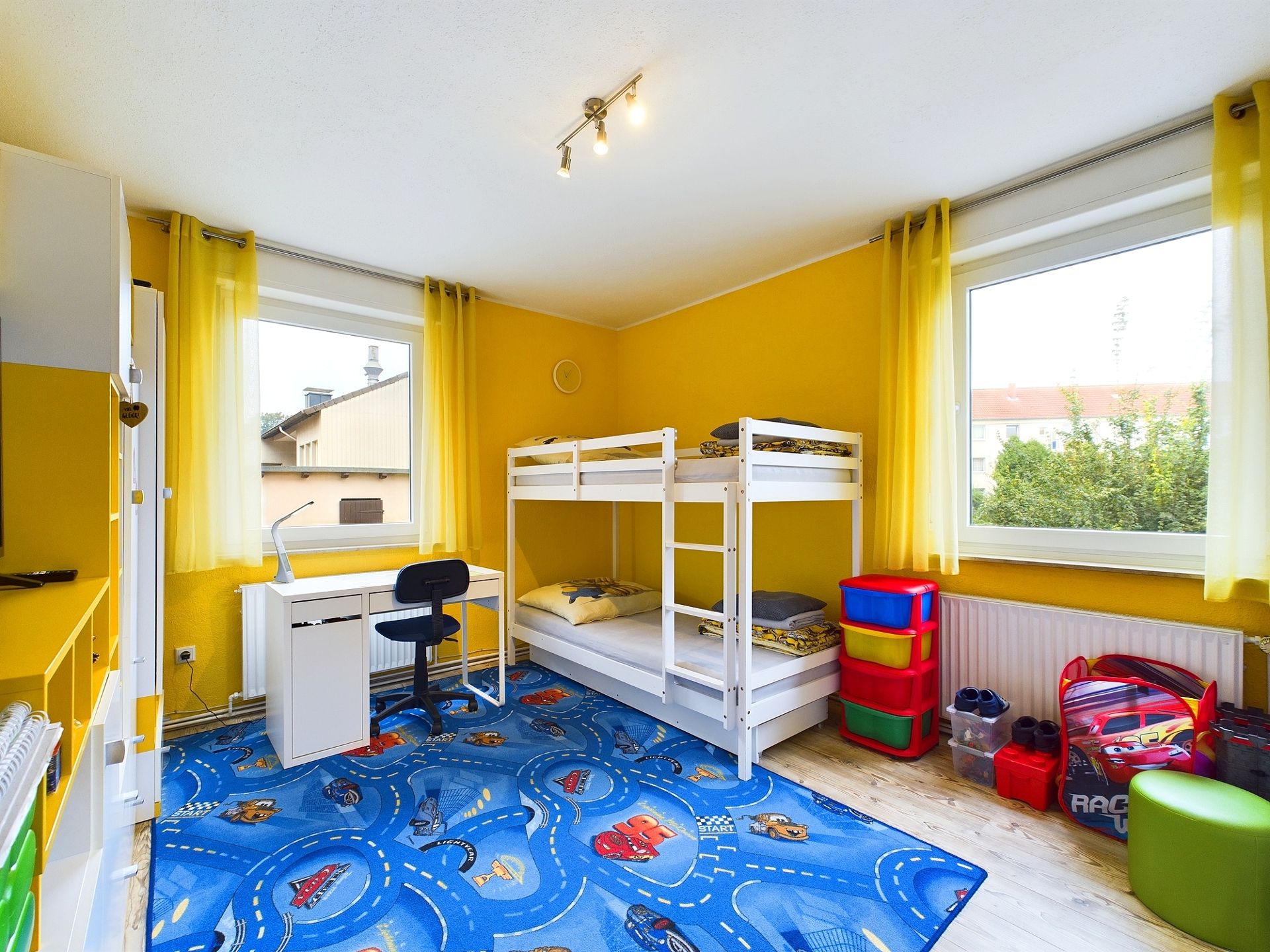 Helles Kinderzimmer mit Etagenbett, buntem Teppich, Schreibtisch und Spielzeugregal.