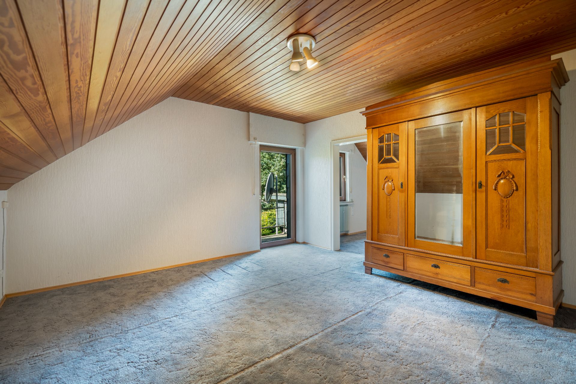 Dachzimmer mit Holzdecke, Teppichboden und großem Holzschrank, Tür zum Balkon.