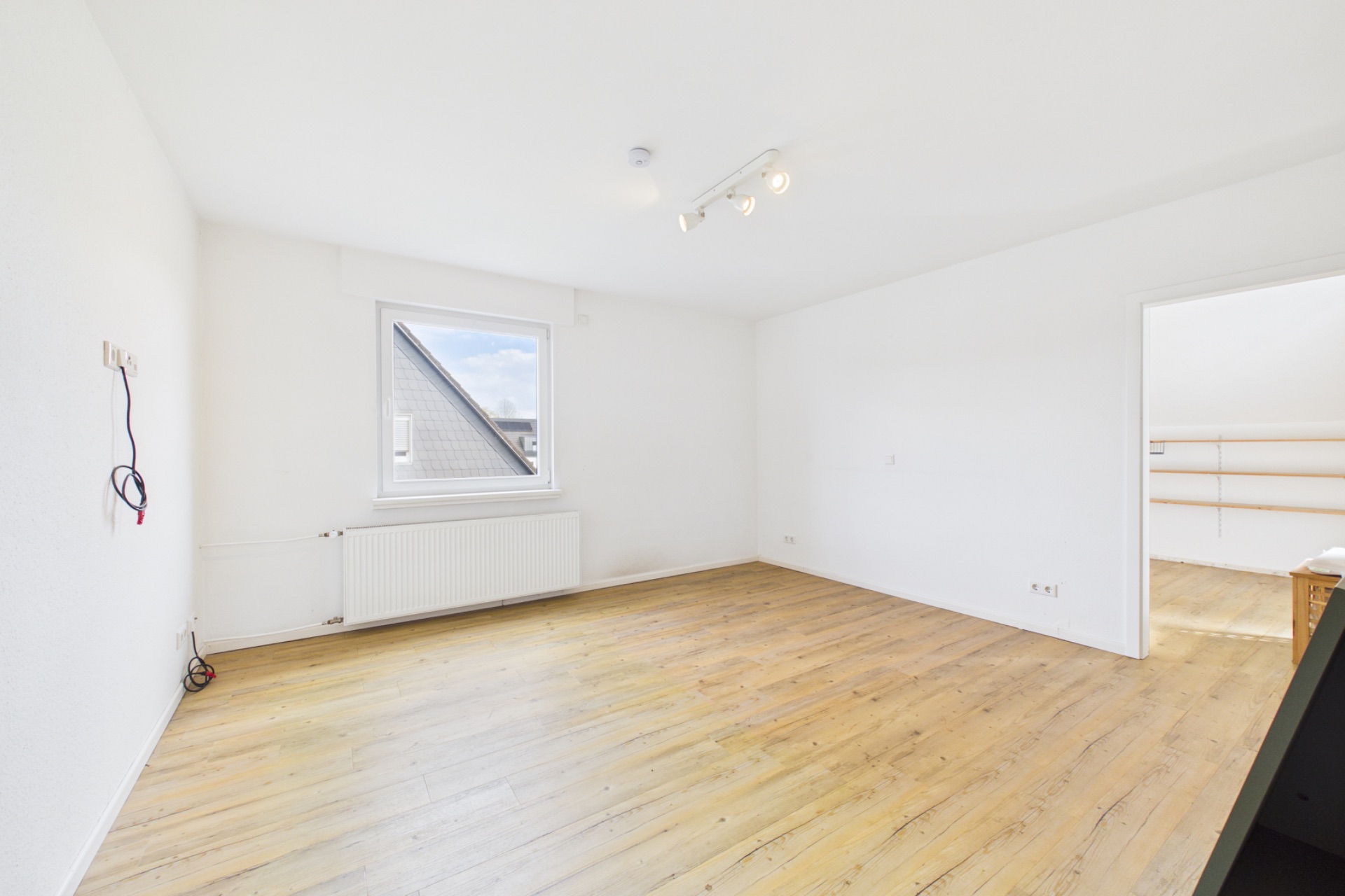 Helles, leeres Zimmer mit Holzfußboden, Fenster, Heizung und Deckenleuchte.