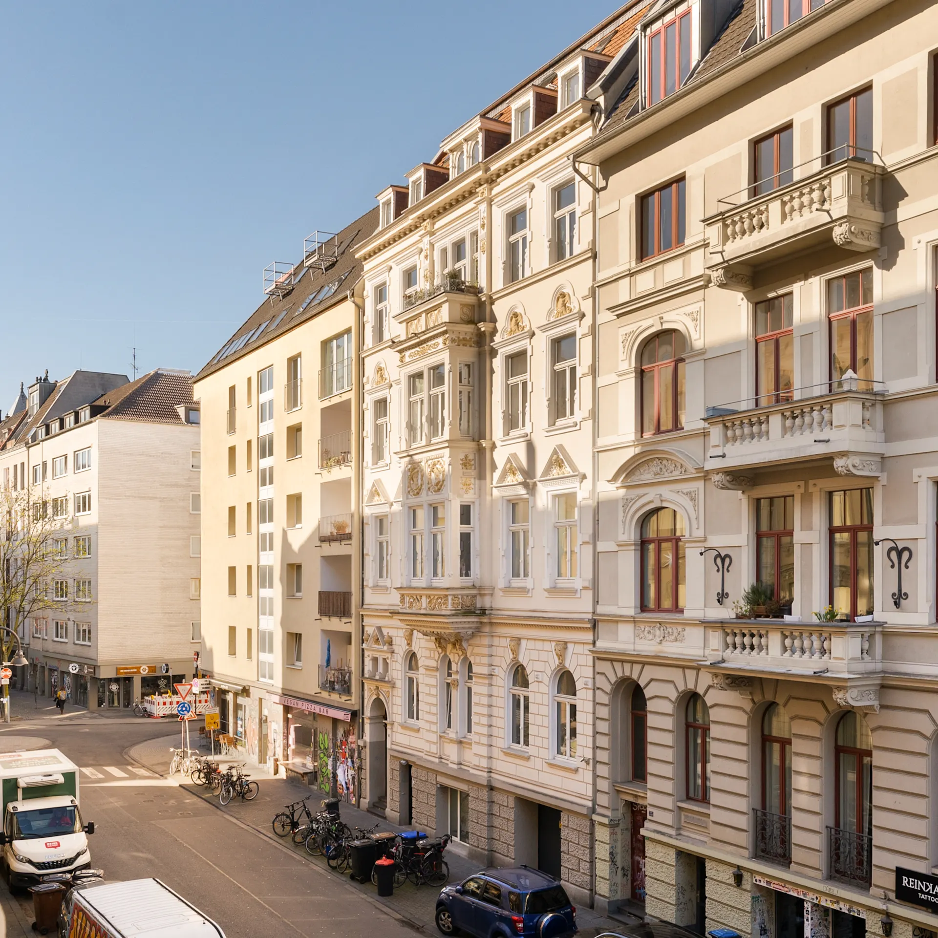 Blick auf eine sonnige Wohnstraße mit mehreren Altbauten, Autos und Fahrrädern am Straßenrand.