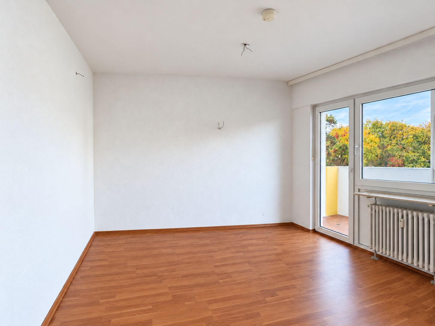 Leerer Raum mit Holzfußboden, weißer Wand, großer Fensterfront und Zugang zum Balkon.