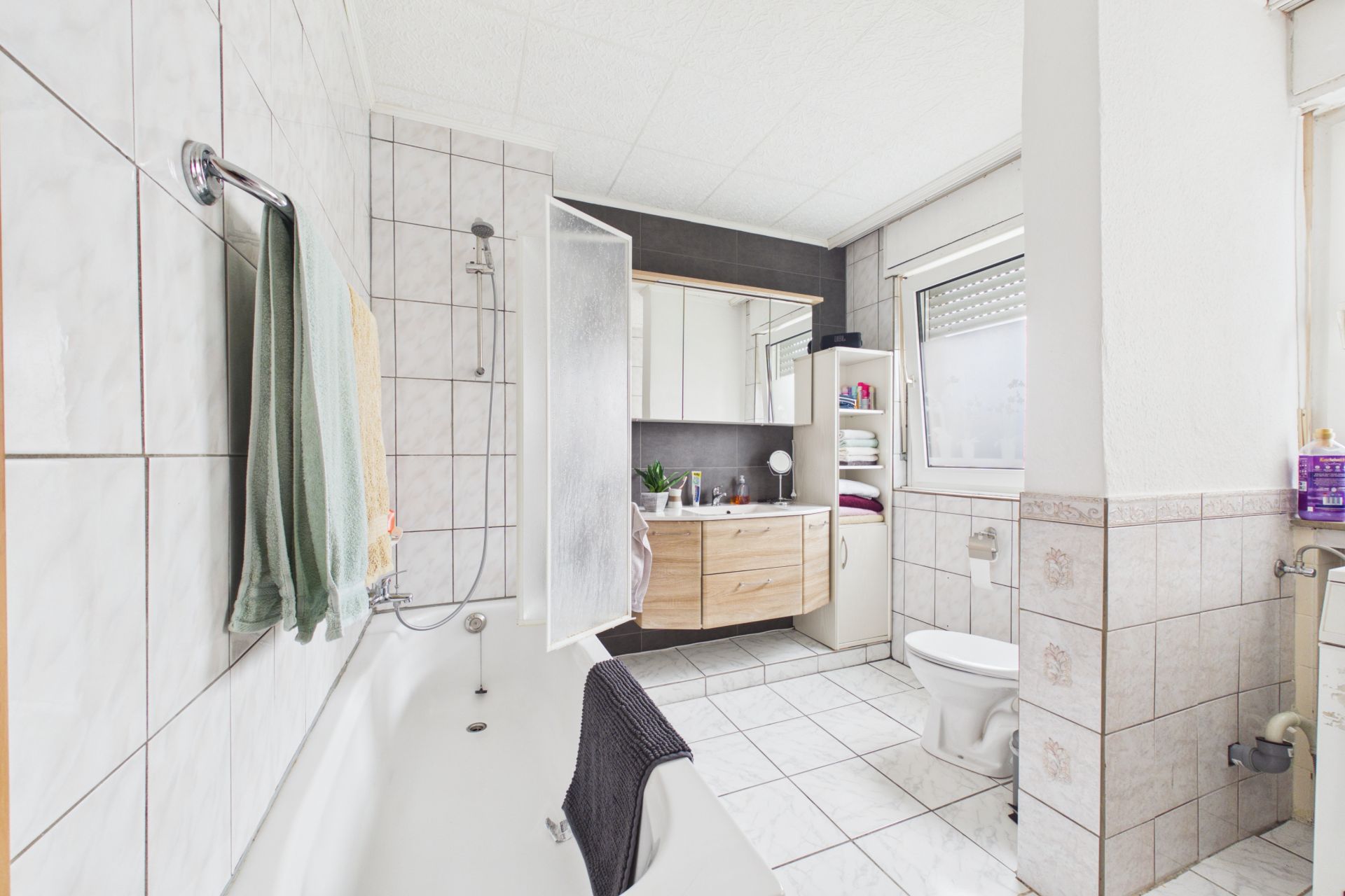 Helles Badezimmer mit Badewanne, Dusche, Waschbecken, WC, Regal und Fenster.