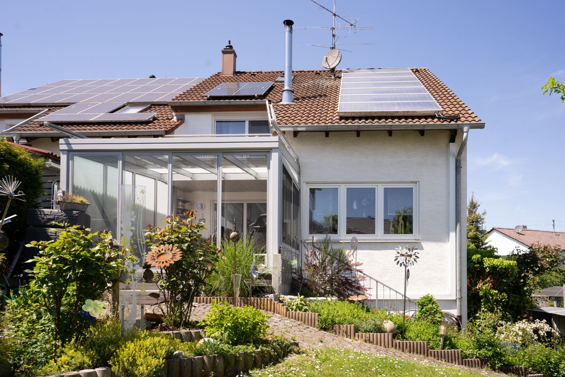 Haus mit Wintergarten, Solarpaneelen auf dem Dach und bepflanztem Garten im Vordergrund.