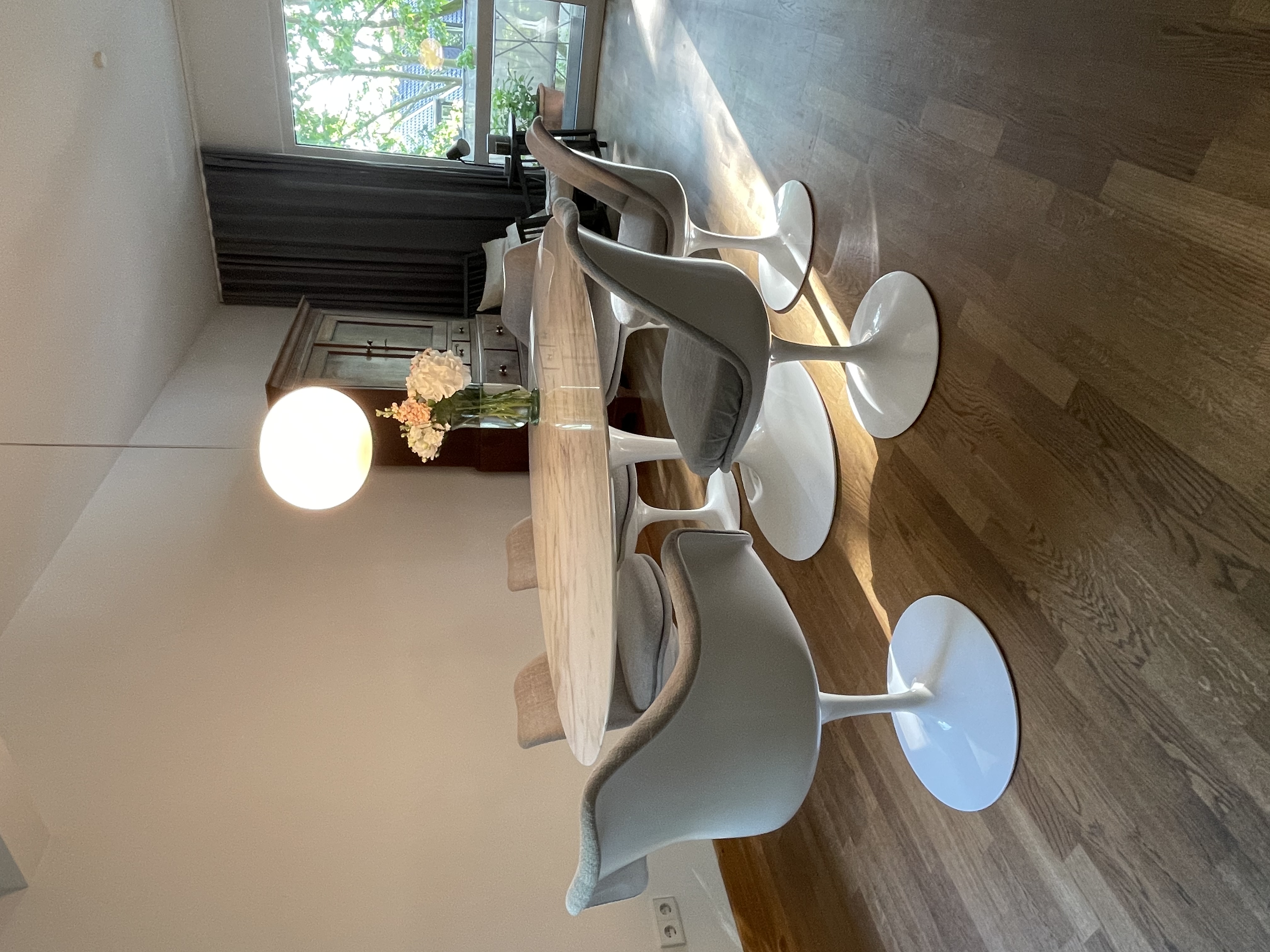 Essbereich mit ovalem Tisch, sechs Stühlen, Blumenstrauß und runder Lampe, Holzboden und Fenster.