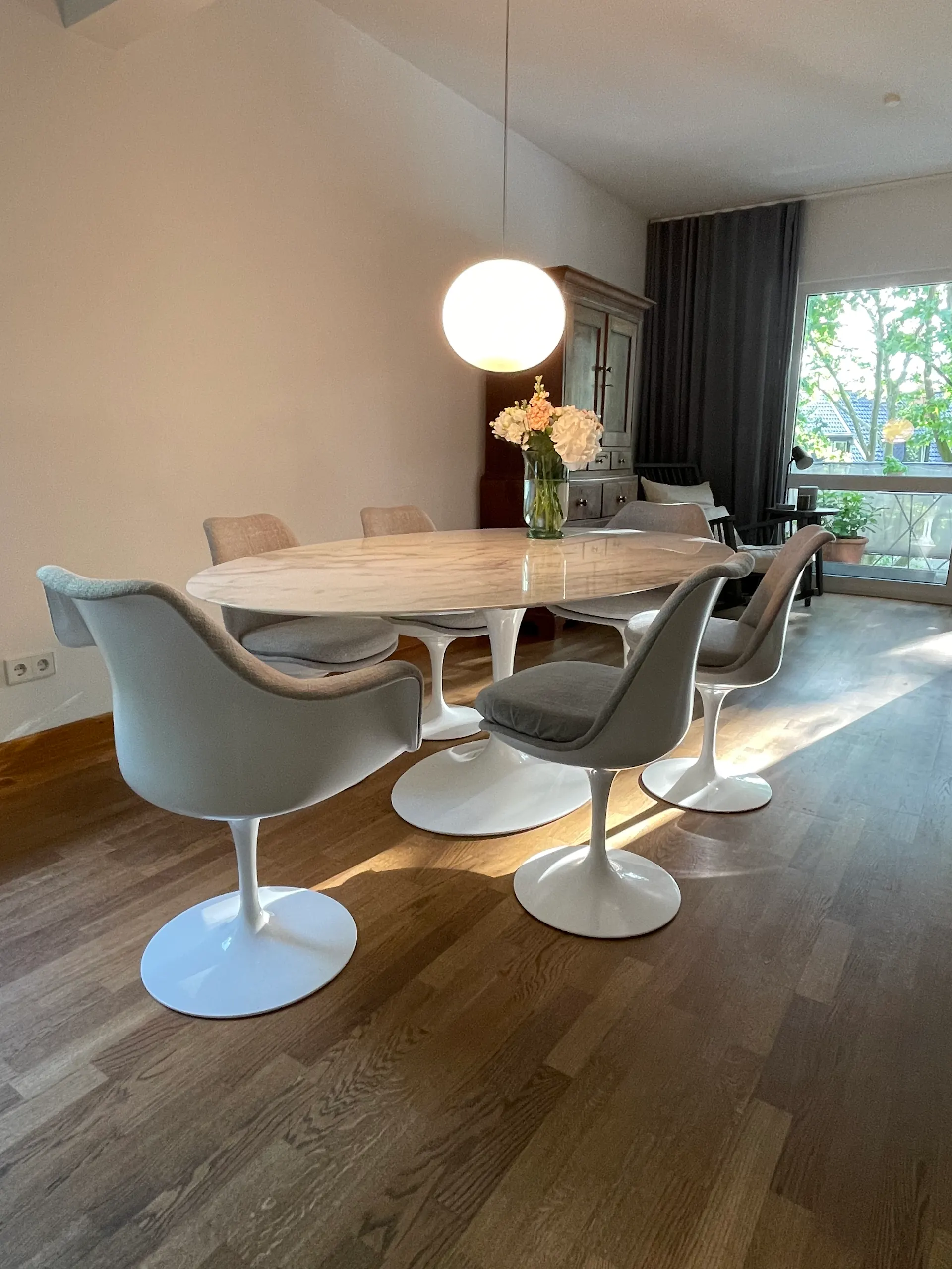 Essbereich mit ovalem Tisch, sechs Stühlen, Blumenstrauß und runder Lampe, Holzboden und Fenster.