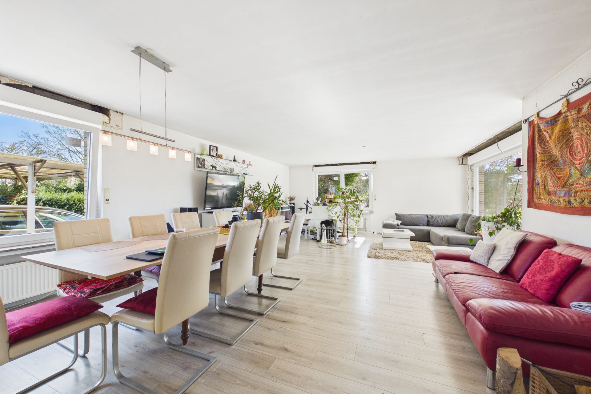 Helles Wohnzimmer mit Essbereich, großem Esstisch, roten und grauen Sofas, Pflanzen und großen Fenstern.