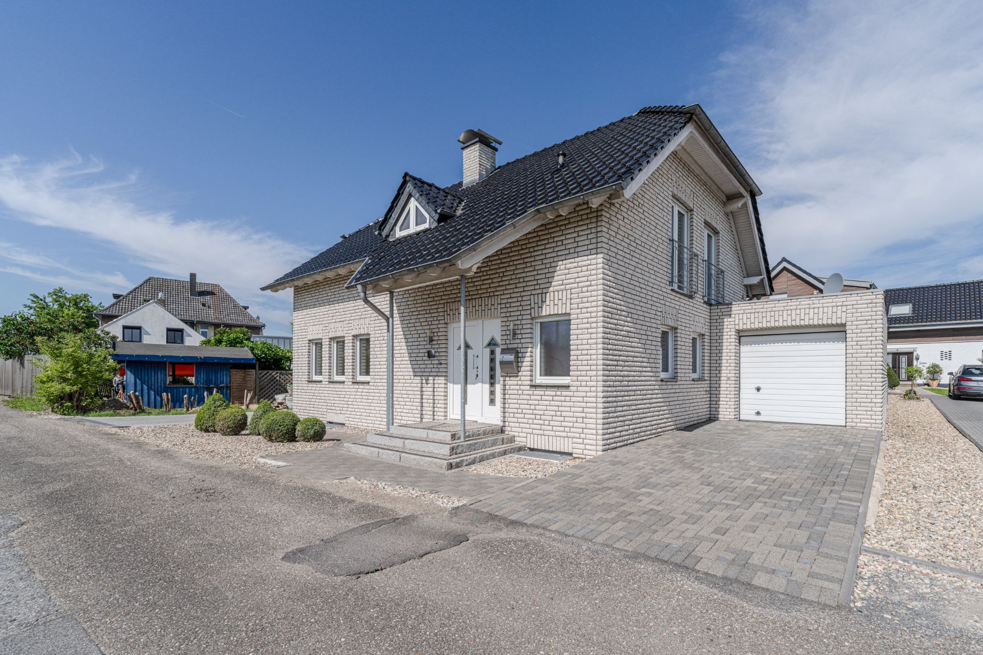 Modernes, zweistöckiges Haus mit hellen Ziegeln, dunklem Dach und angegliederter Garage an einer ruhigen Straße.