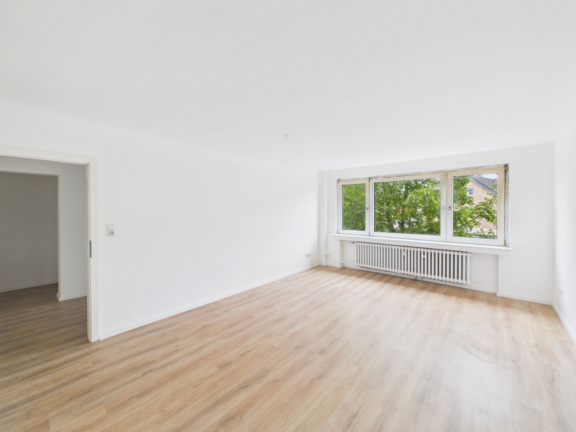 Helles, leeres Wohnzimmer mit Holzfußboden und großem Fenster mit Blick auf Bäume.