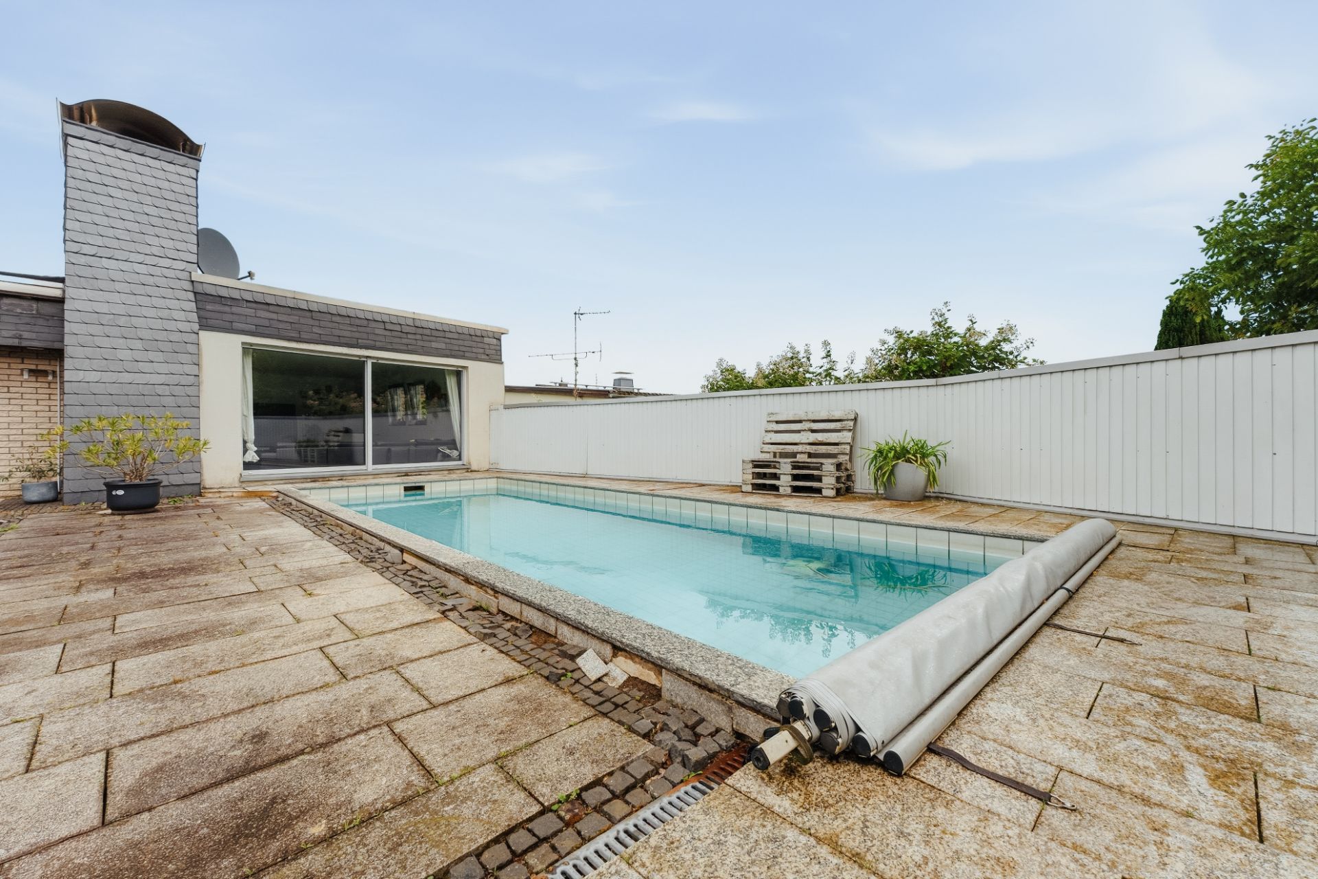 Rechteckiger Außenpool mit Steinboden, angrenzender Terrasse und weißen Zaun im Hintergrund.