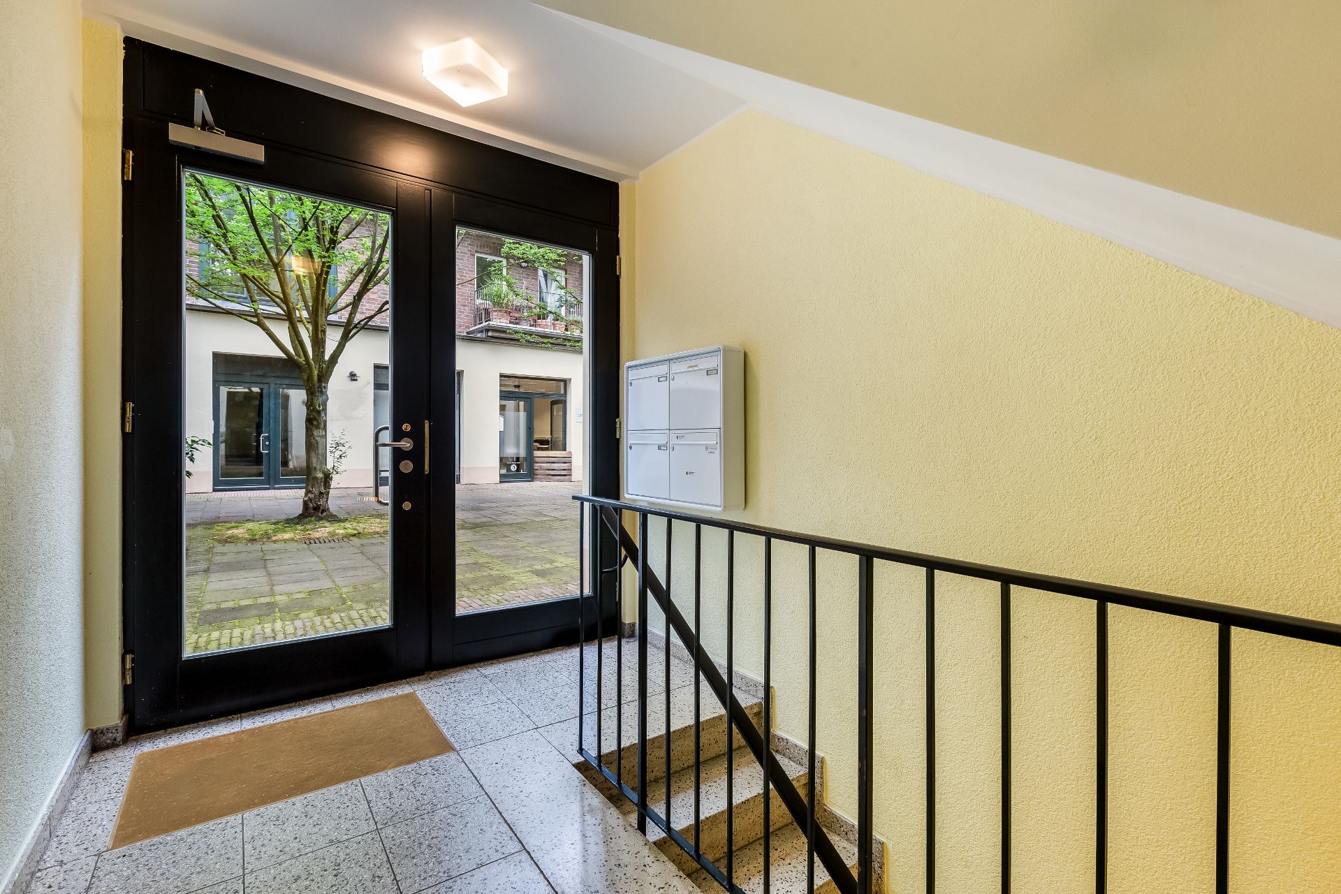 Eingangsbereich mit Glastür, Briefkästen und Treppe, Blick auf Innenhof mit Baum.