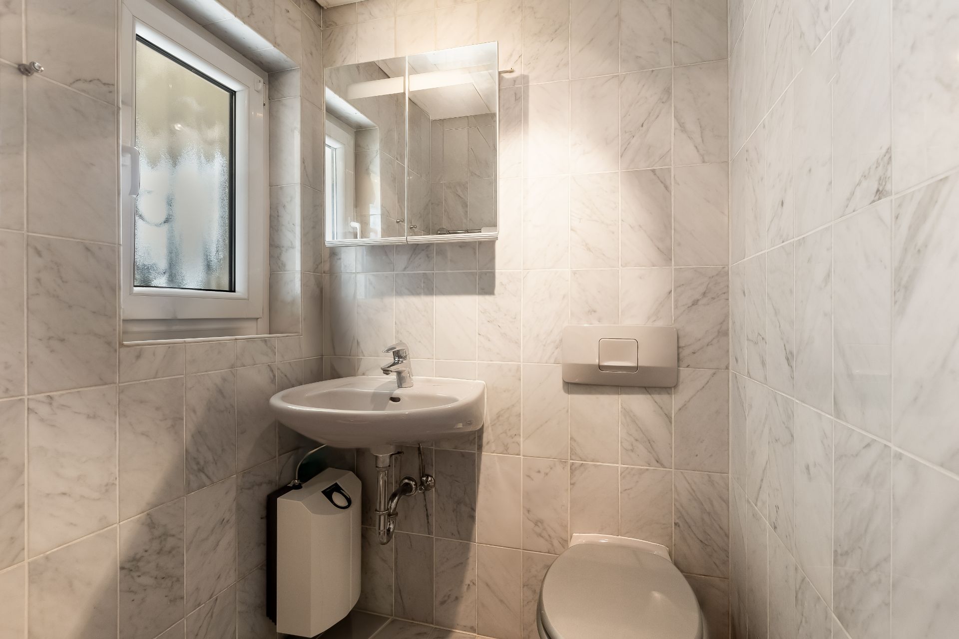 Kleines Badezimmer mit Waschbecken, Spiegel, Fenster und Toilette mit Fliesenwänden.
