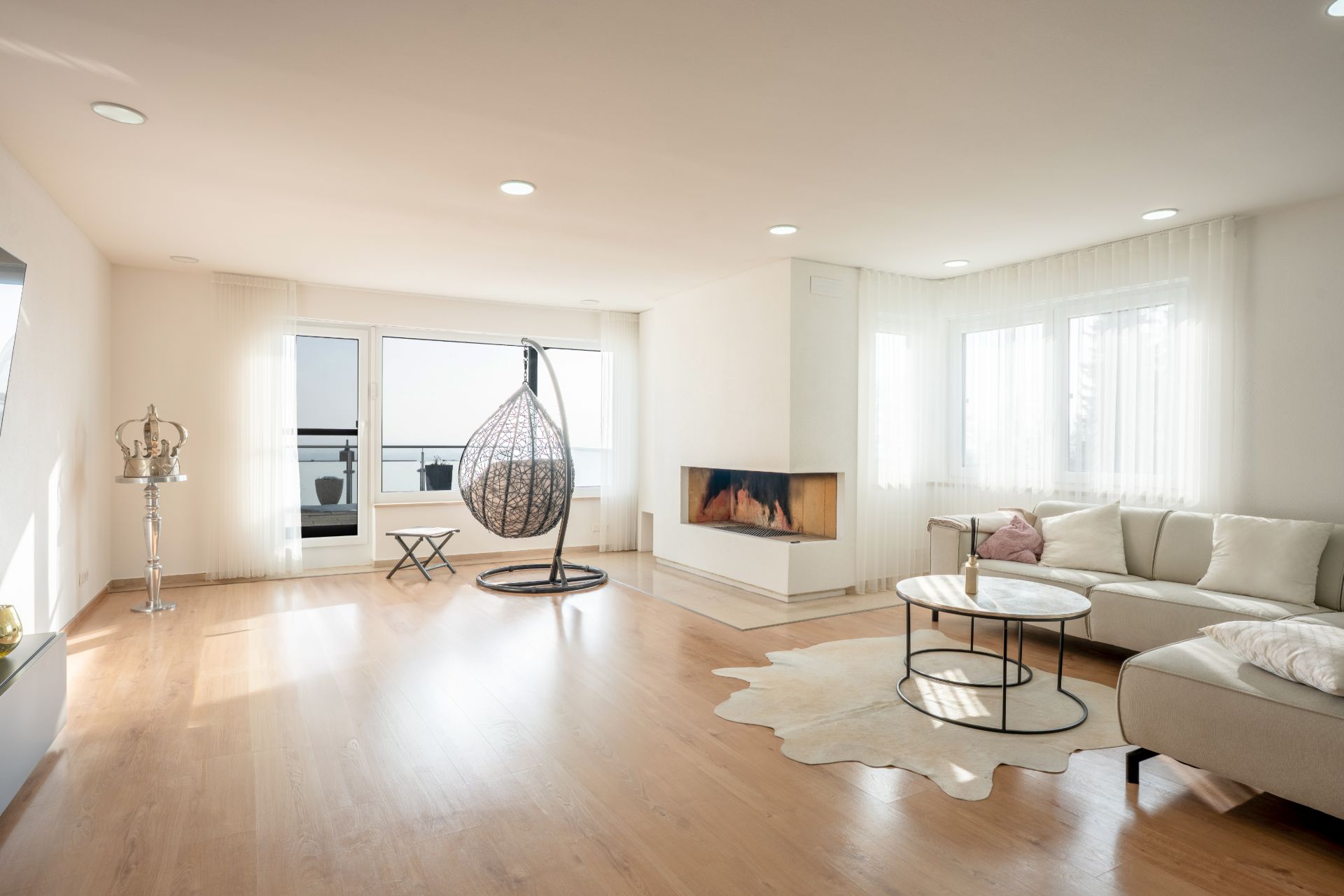 Helles Wohnzimmer mit Ecksofa, Kamin, runder Hängeschaukel und Couchtischen auf Holzfußboden.